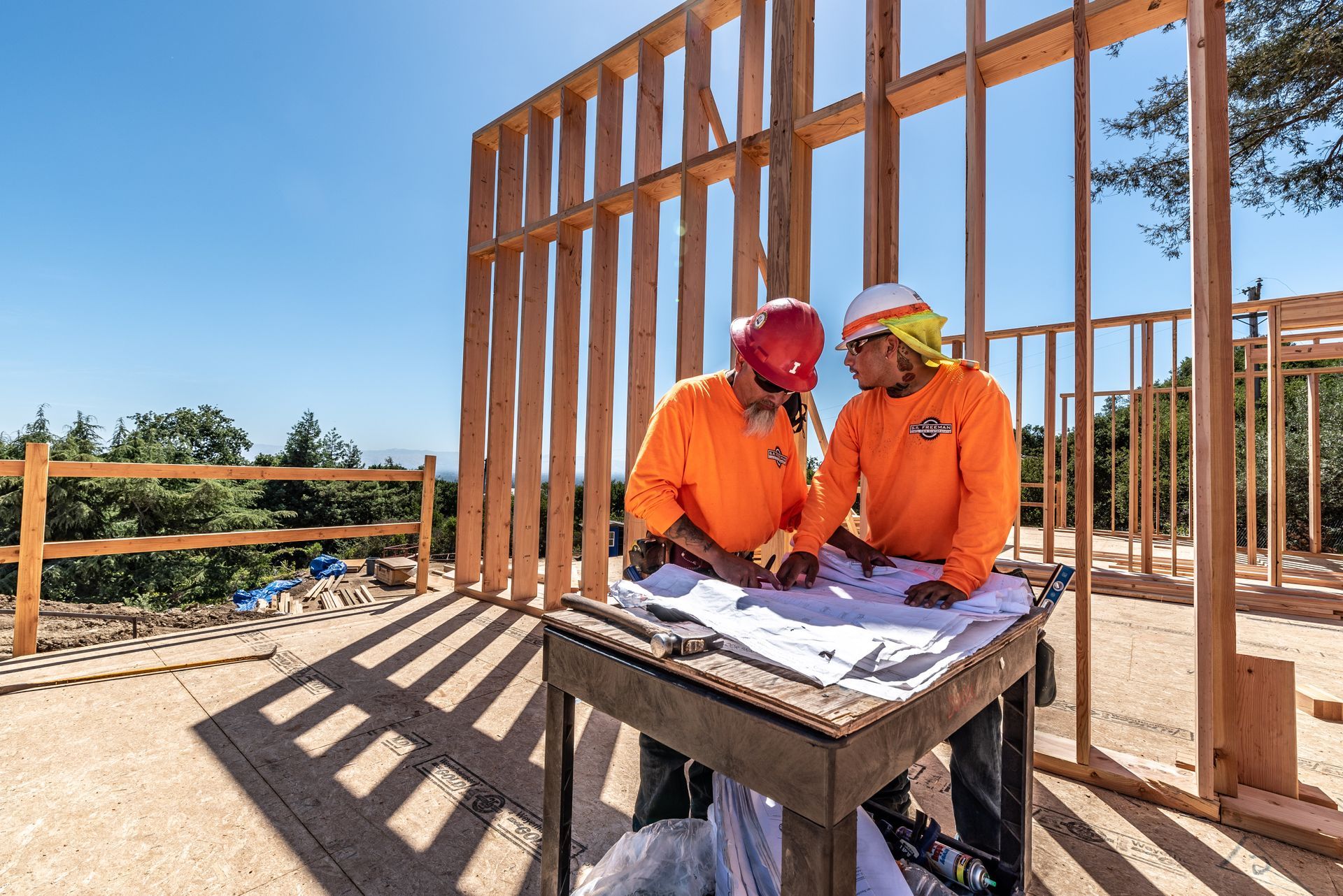 Two construction workers in orange vests review plans at a house frame under construction.