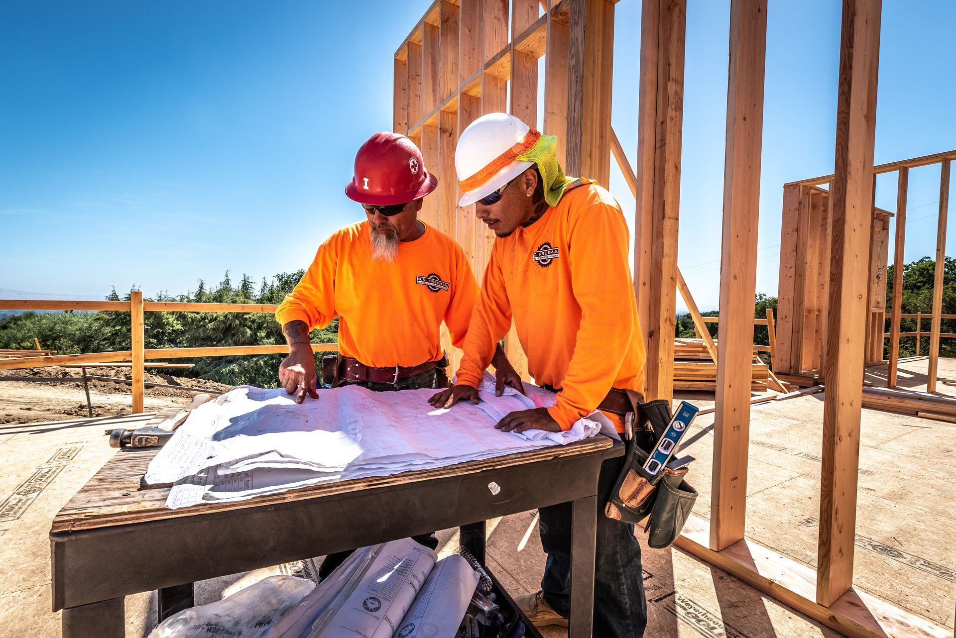 Two construction workers in orange vests review blueprints at a framing site.