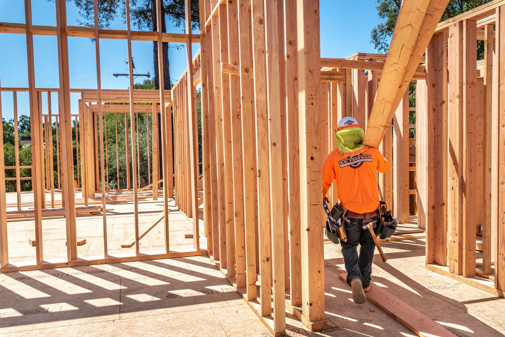 Worker in orange shirt framing a house under construction, carrying tools inside a wooden structure.