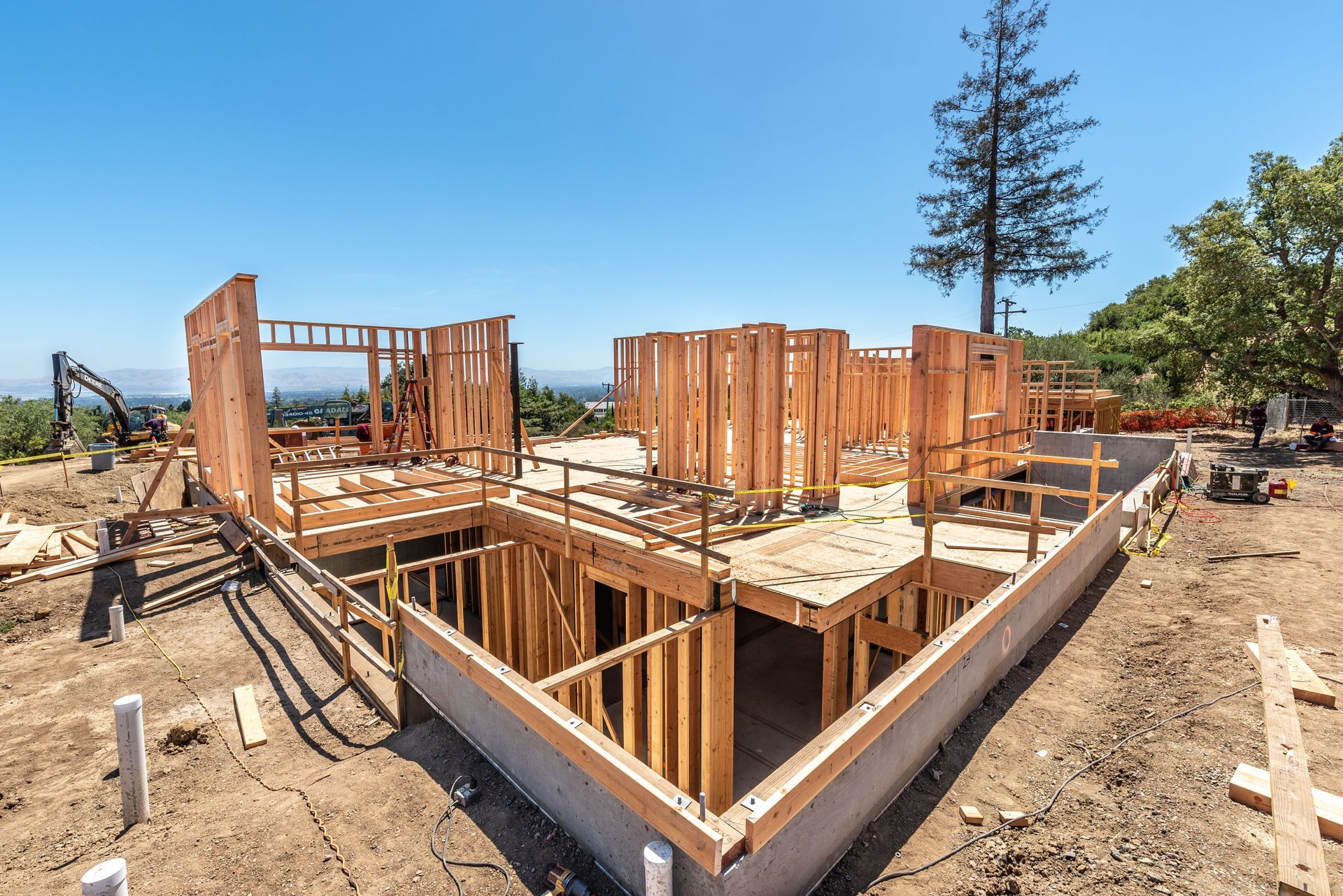 Foundation and wood framing of a house under construction on a sunny, open lot with trees and blue sky.
