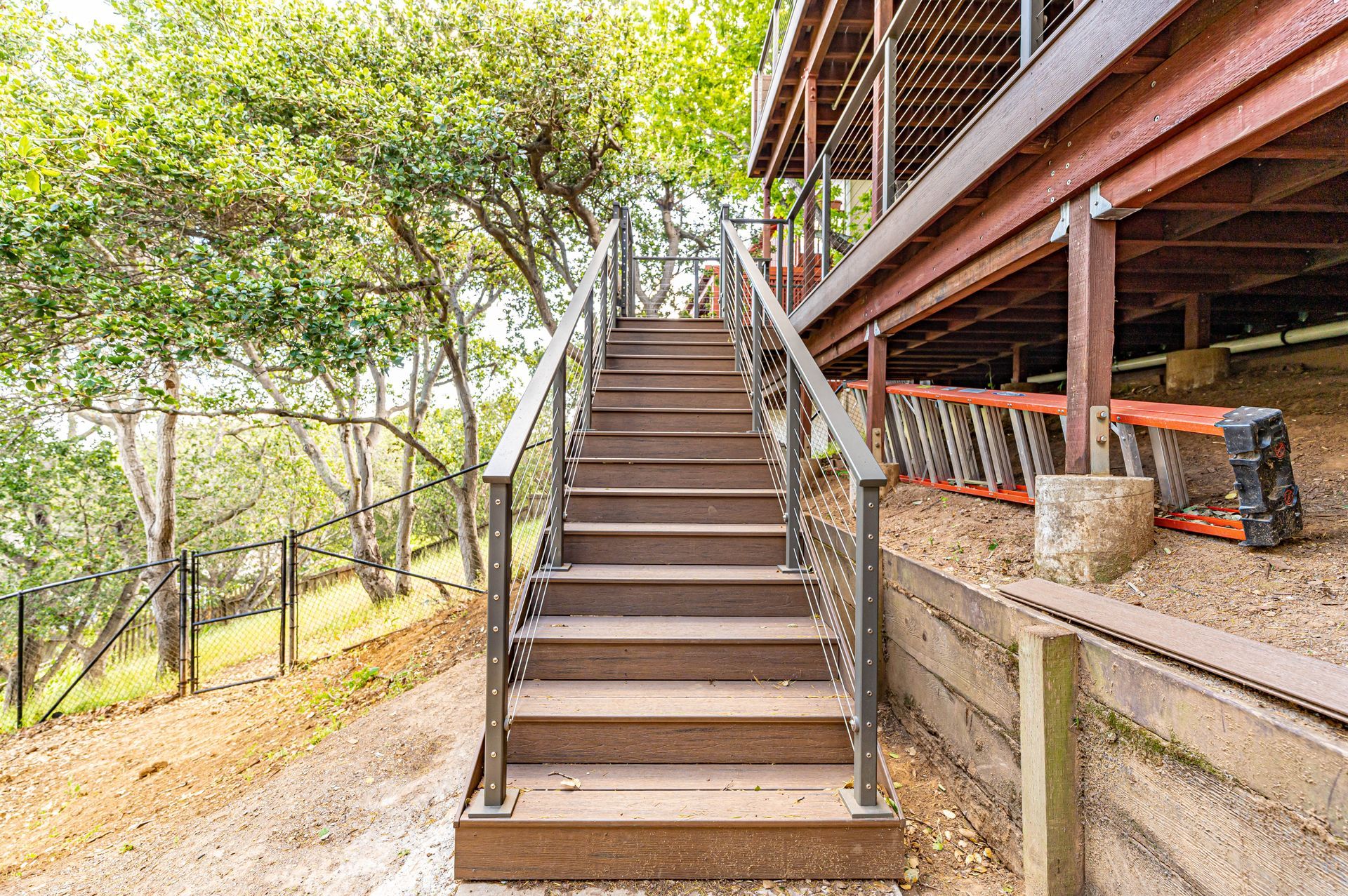 Outdoor wooden staircase leading to an elevated deck beside trees and a concrete retaining wall