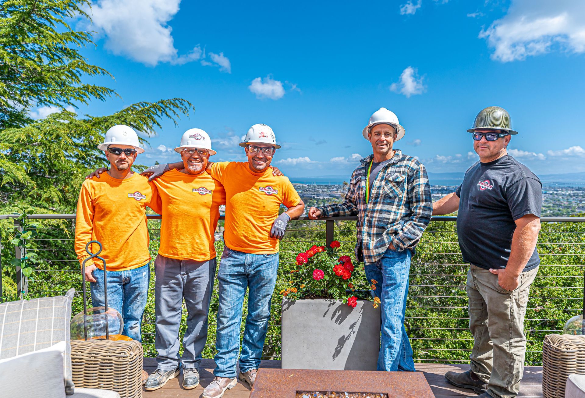 Five workers in hard hats standing outdoors by a planter with red flowers and a city view behind them