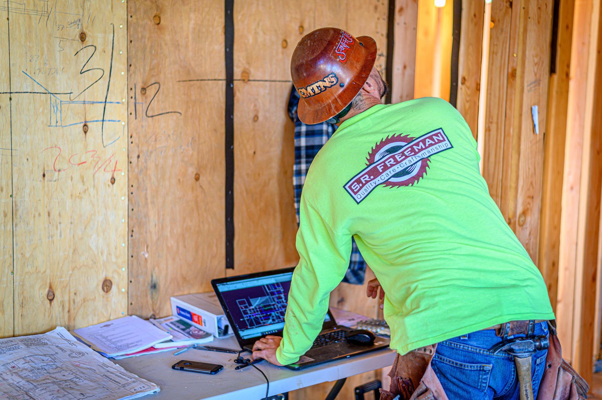 Worker in neon green shirt and cowboy hat using a laptop at a wooden workbench inside a framed unfinished room