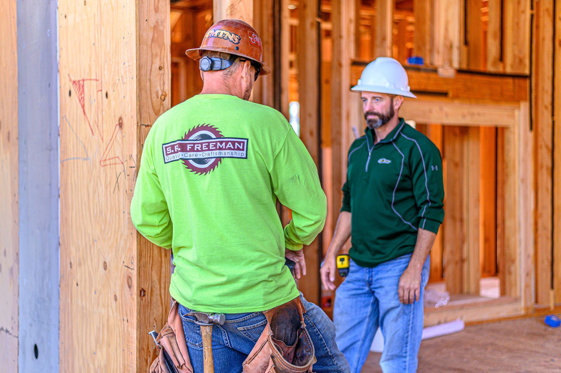Two construction workers talking at a wood-framed building site, one in a green shirt and one in a white hard hat.