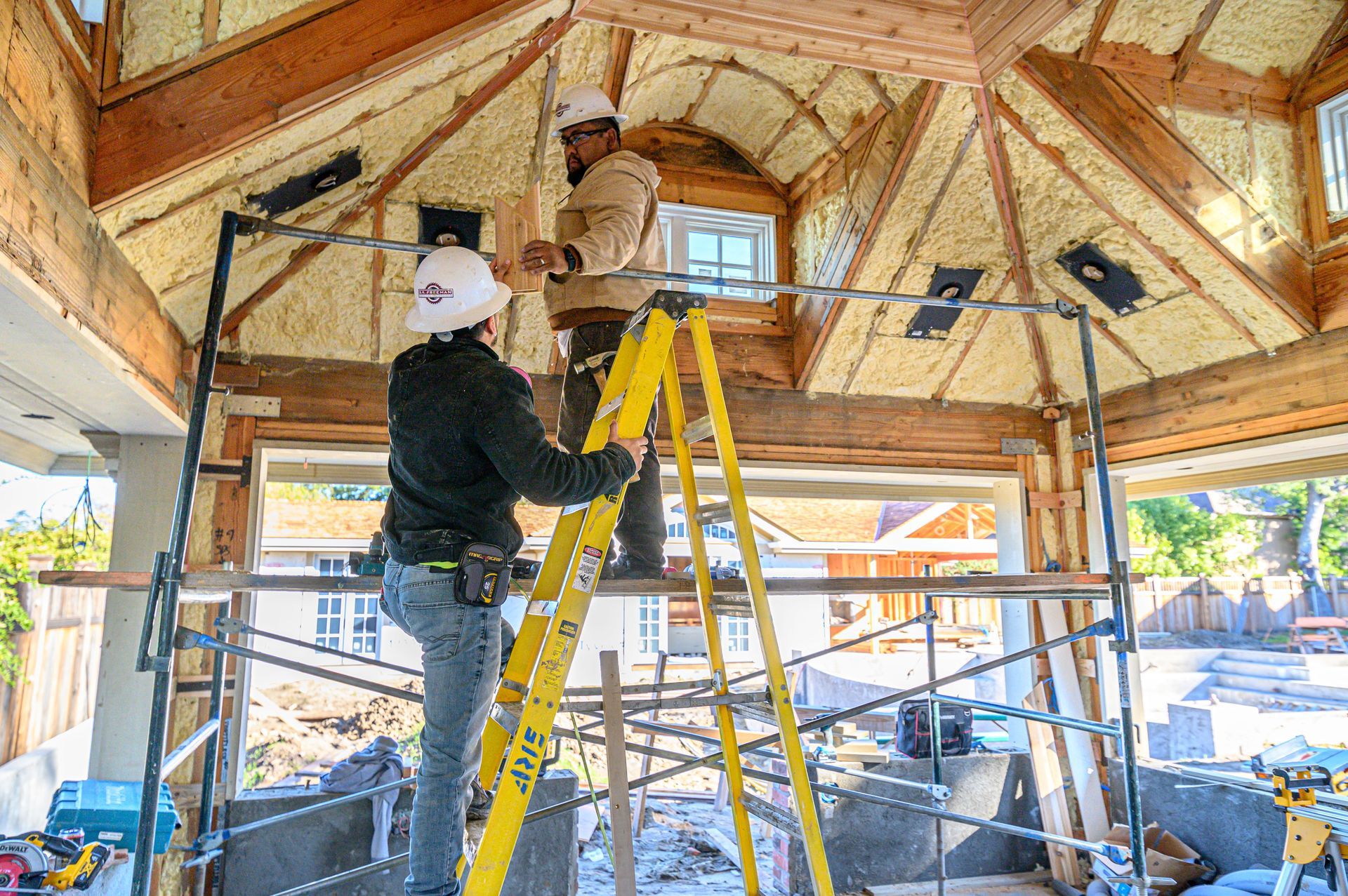 Workers in a scaffolded attic, installing insulation under a peaked wooden ceiling.