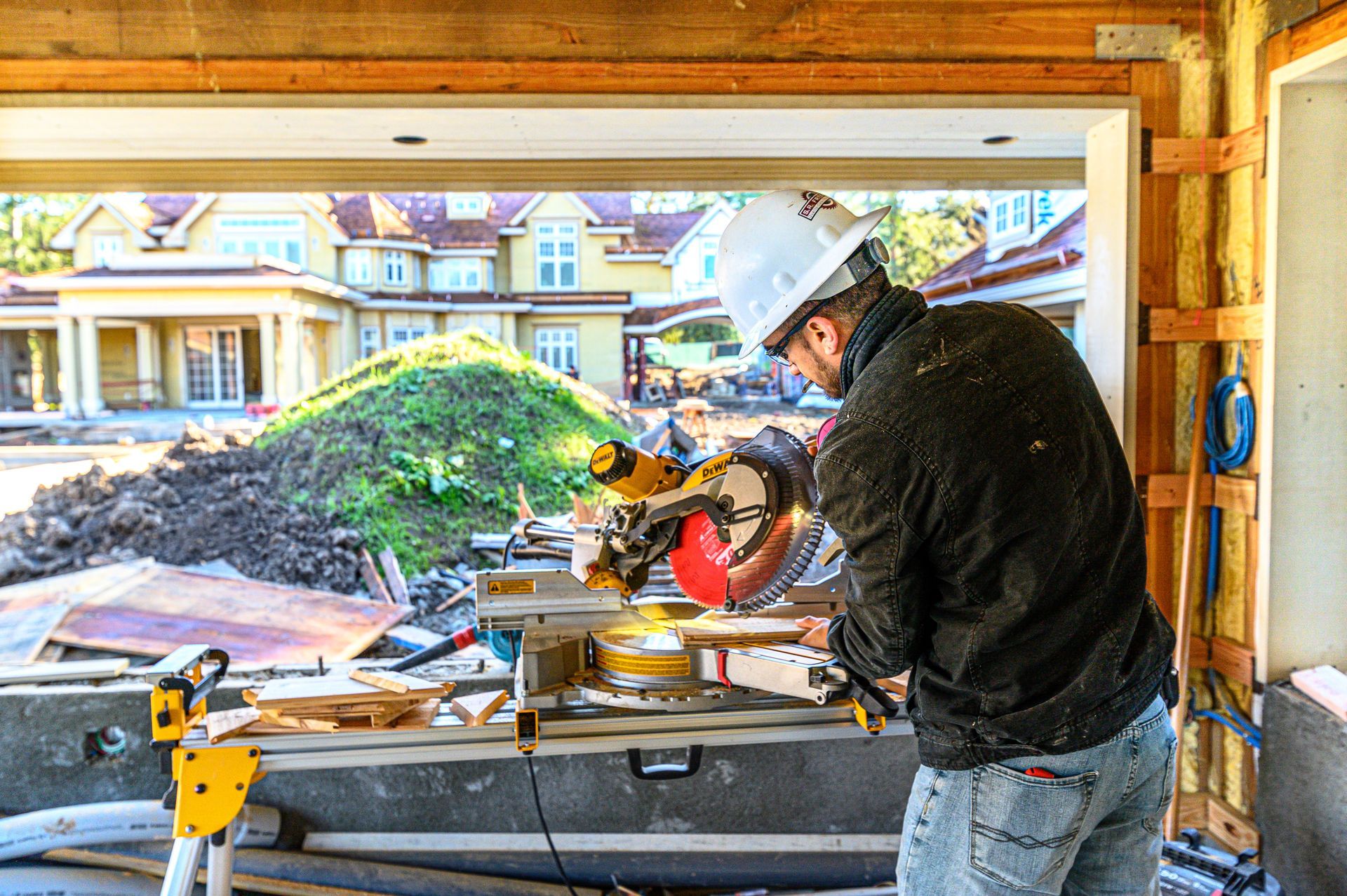 Construction worker cutting wood in a garage with a miter saw, wearing a hard hat and earmuffs