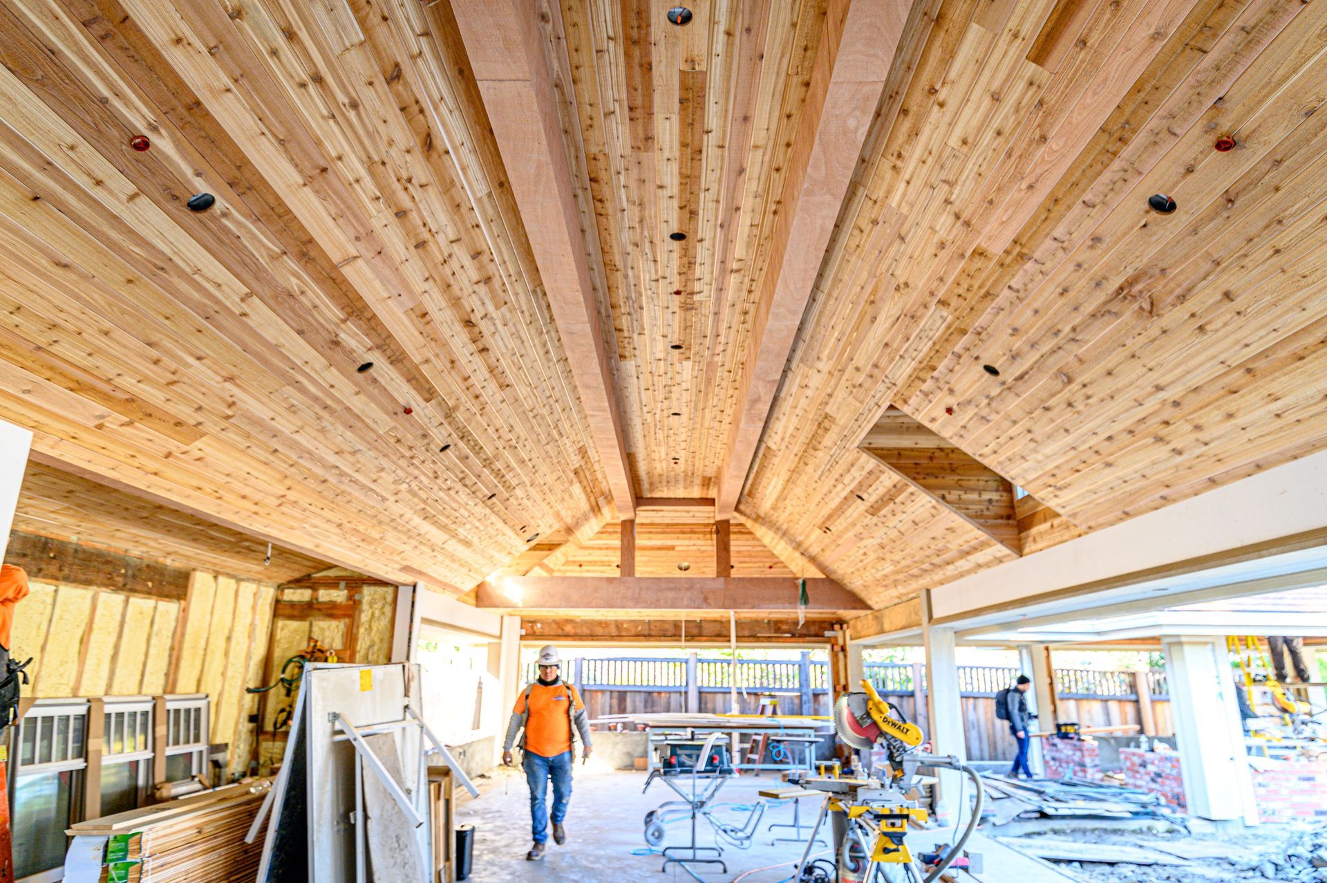 Wood-paneled interior under construction with workers, tools, and exposed beams in a bright open space