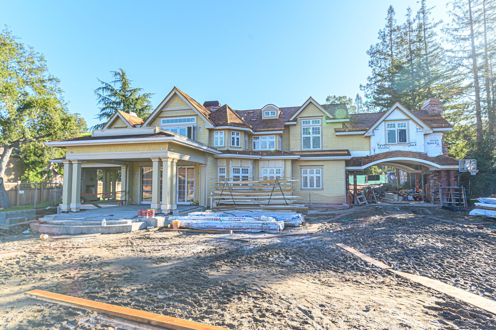 Large stone house under construction with a covered porch, bare yard, and tall trees around it.