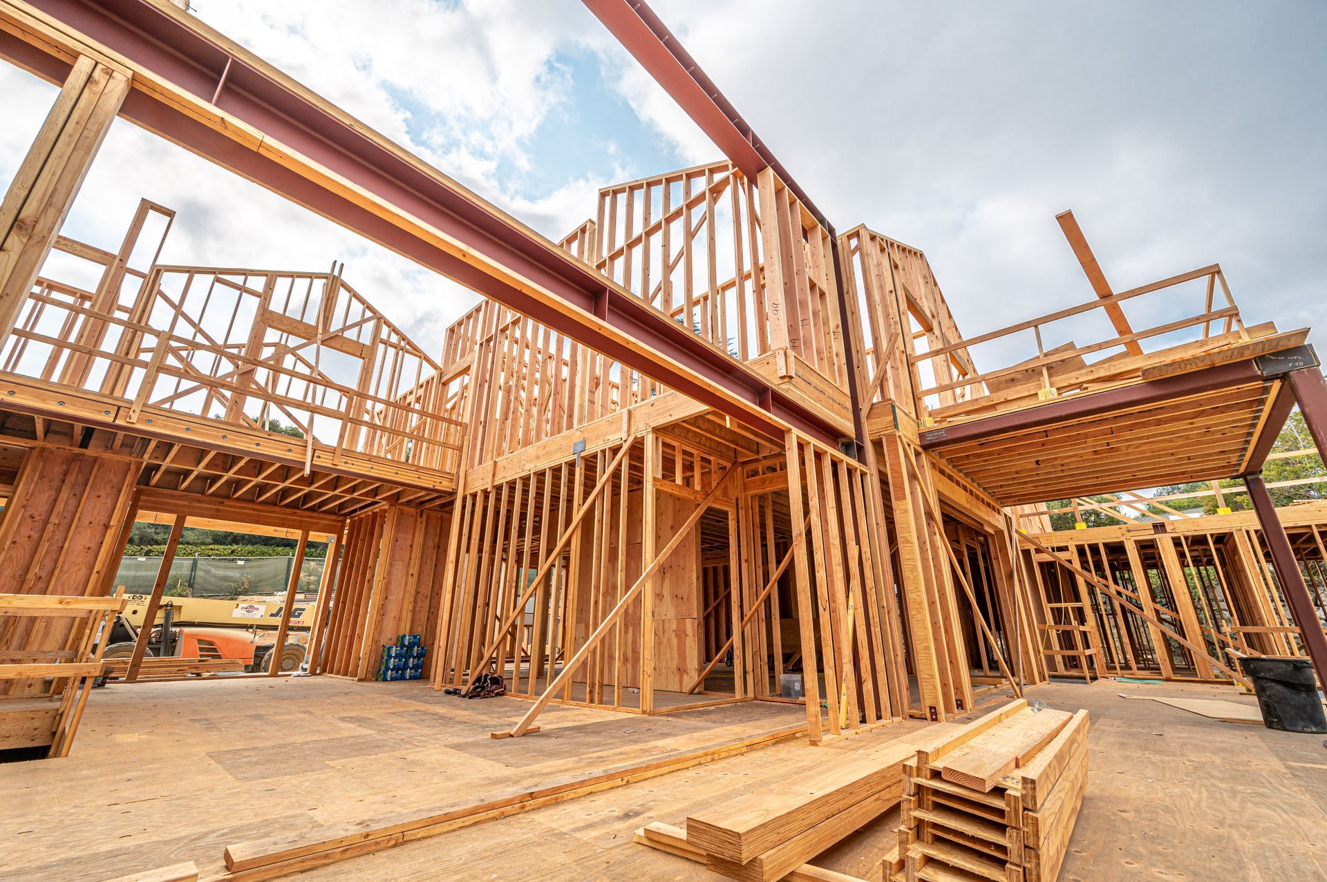 Wood-framed house under construction with exposed beams, scaffolding, and stacks of lumber under a blue sky