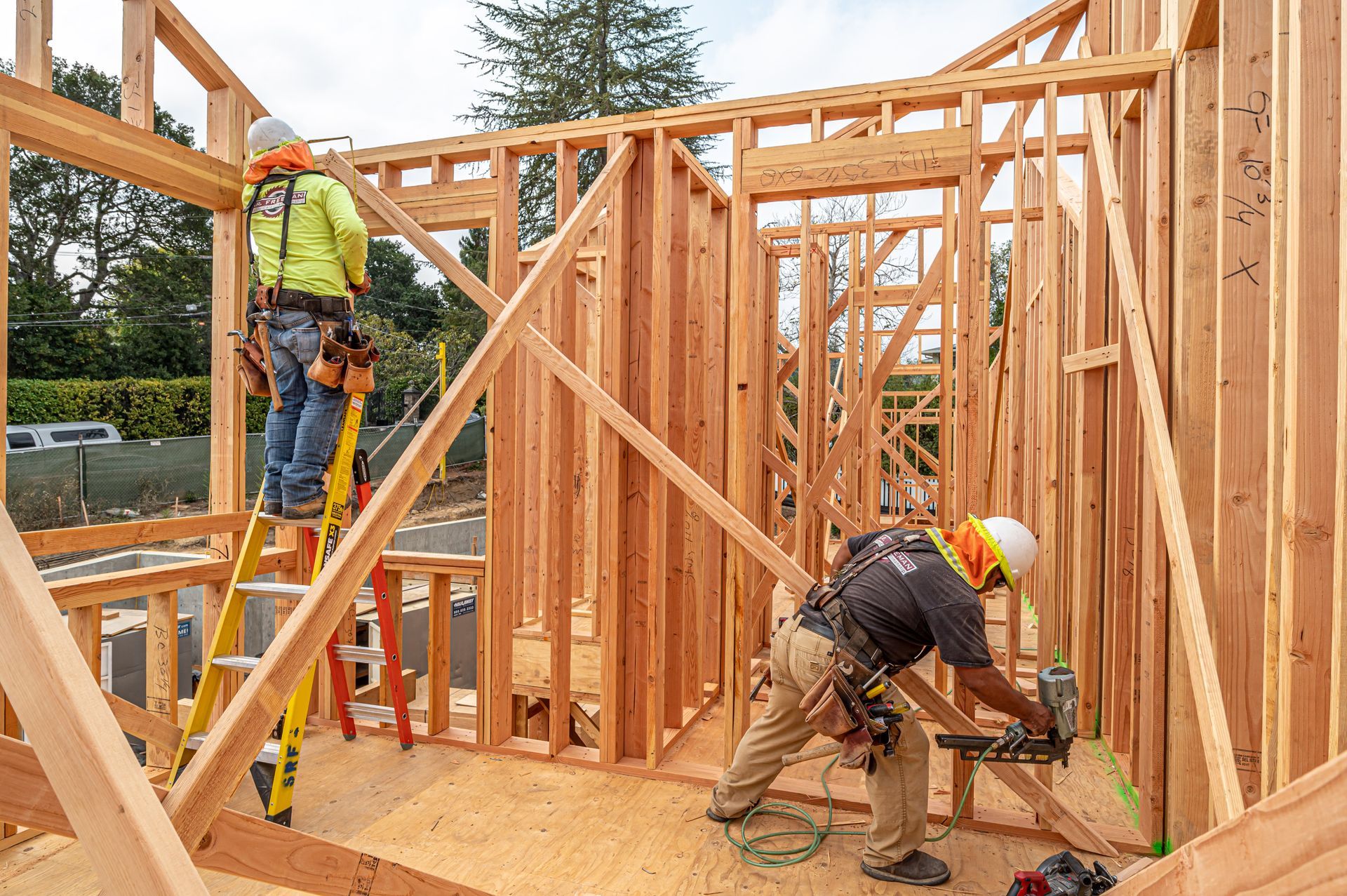 Construction workers framing a wooden house, one using a saw while another stands on the wall frame.