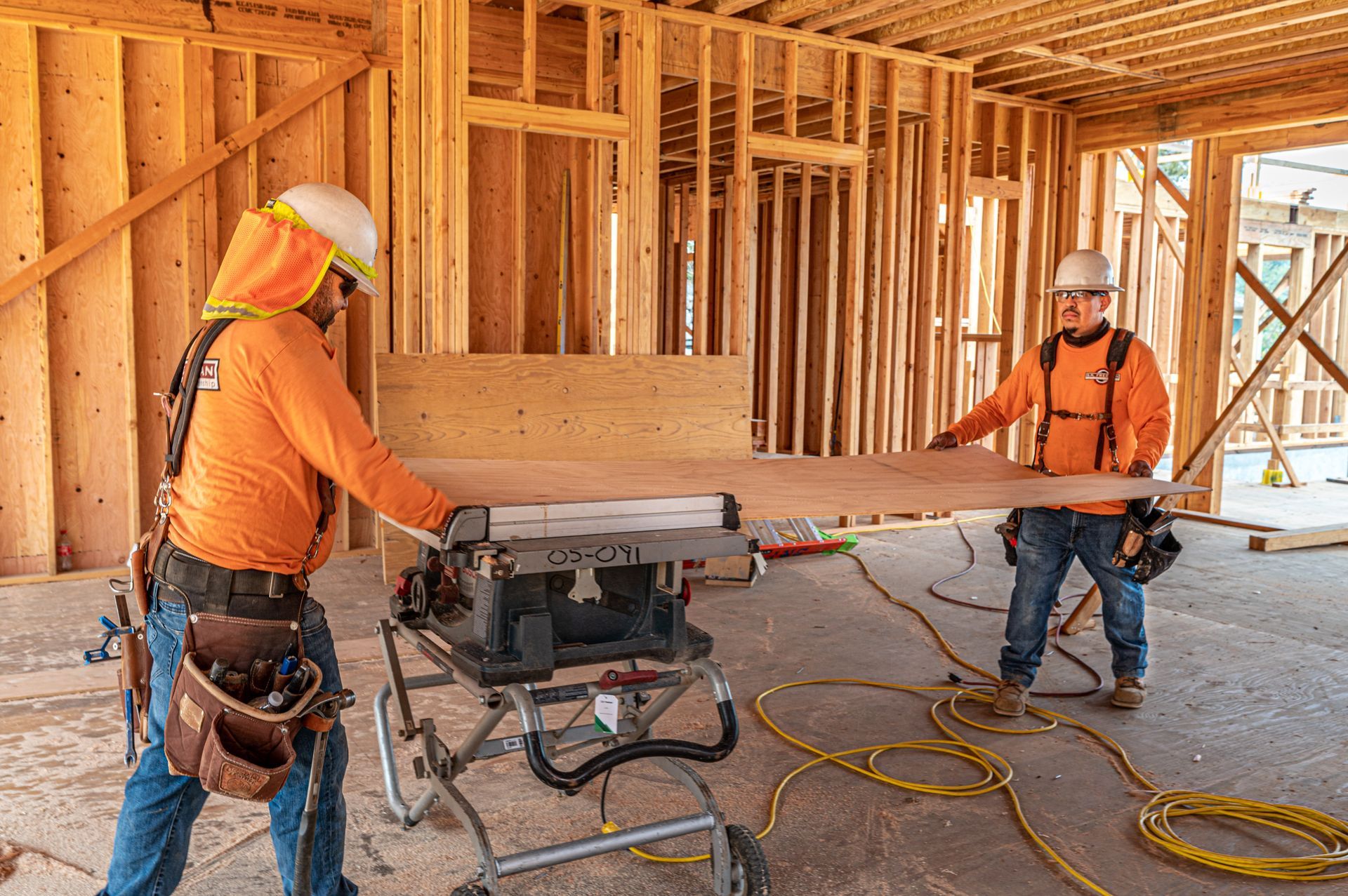 Two construction workers in orange shirts operate tools inside a wood-framed building under construction