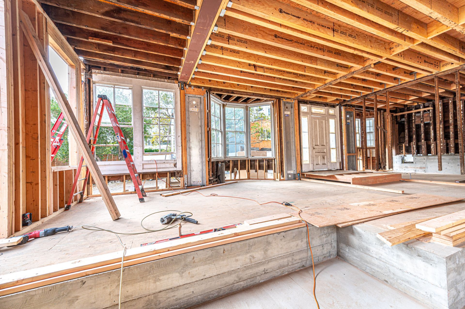 Interior of a house under renovation, with exposed wooden framing, windows, and a ladder on the floor.