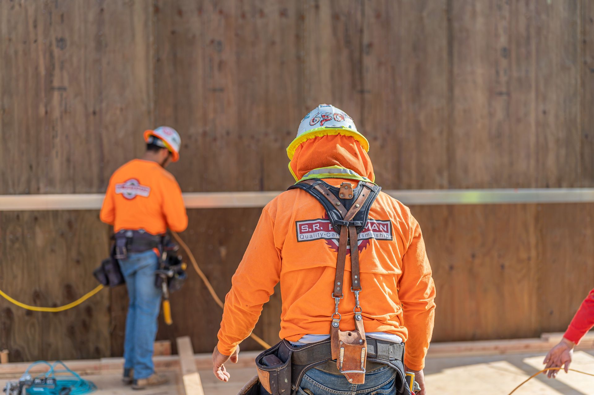 Two workers in orange safety gear standing on a construction site with a large wooden wall.
