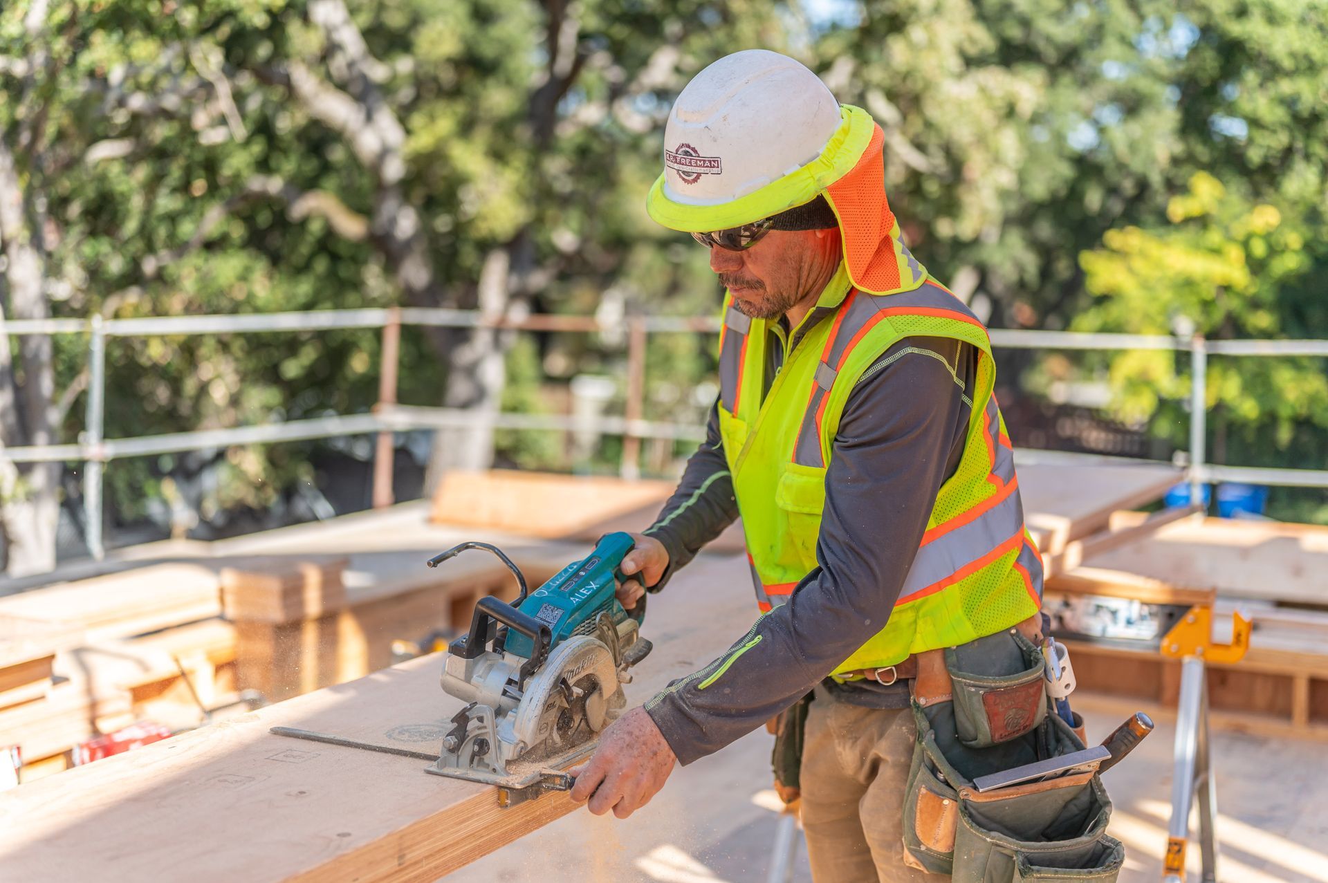 Construction worker cutting wood on a rooftop with a circular saw, wearing a hard hat and safety vest.