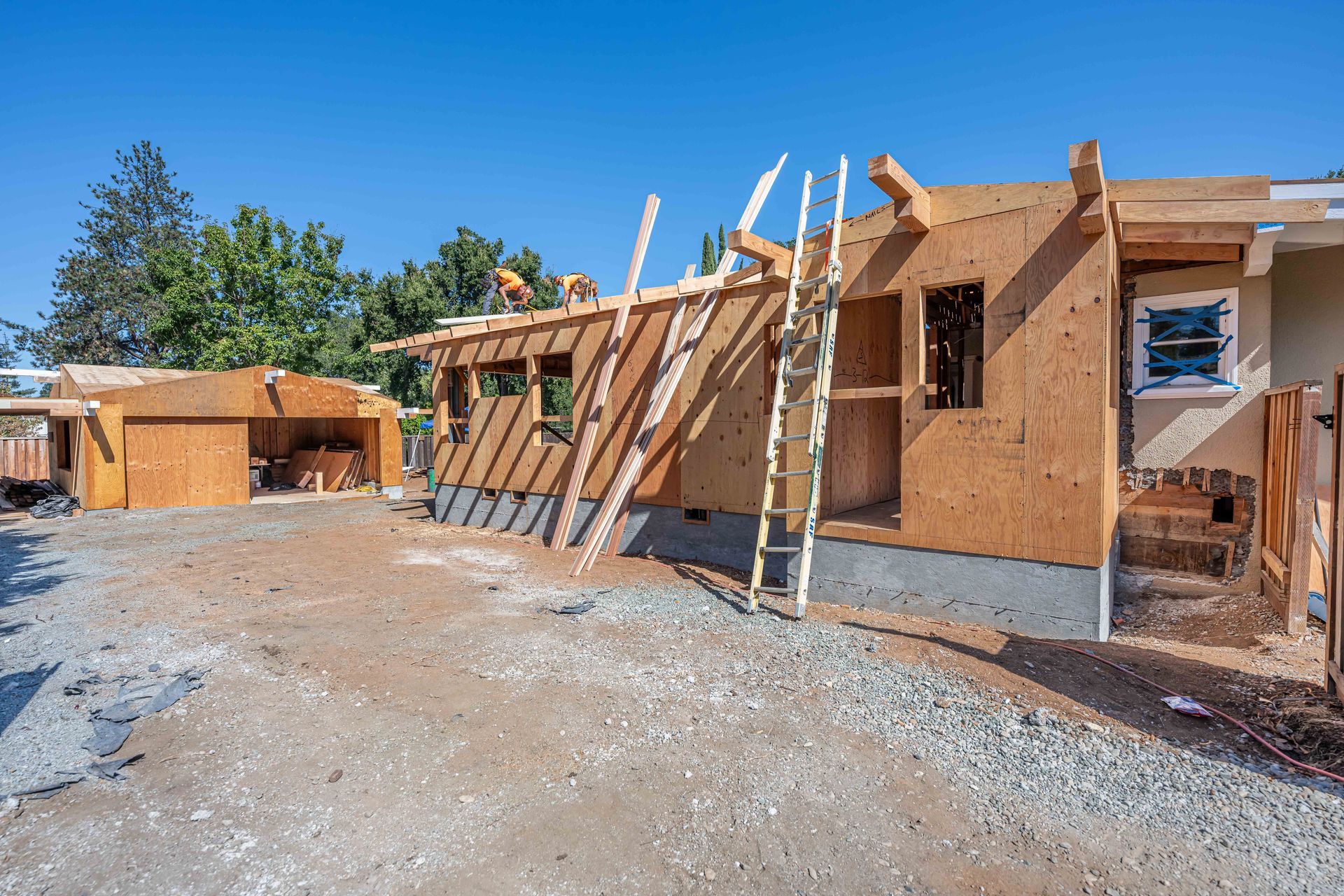 Partially built wooden house framing on a sunny construction site with dirt ground and blue sky