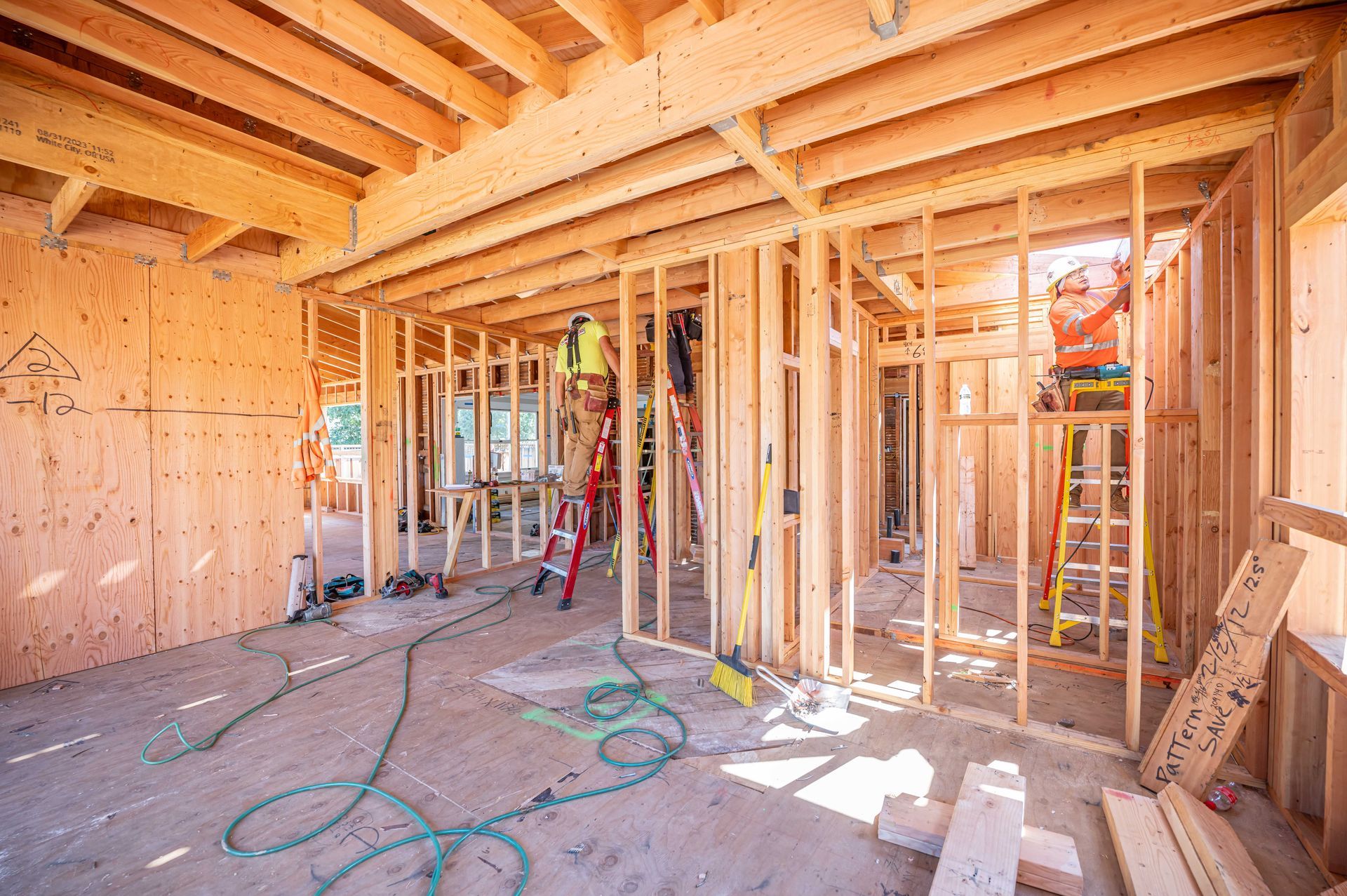 Unfinished house interior with exposed wooden framing, ladders, and construction tools in sunlight
