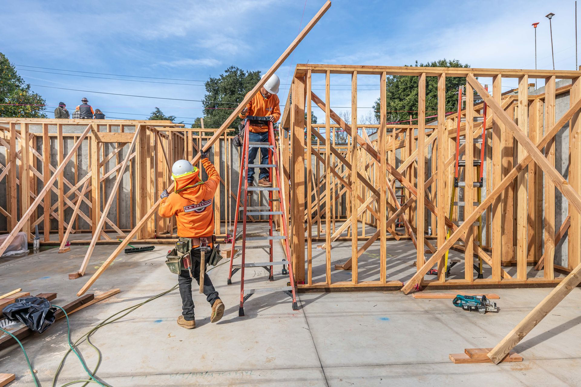 Construction workers framing a house, with wooden walls, ladders, and a concrete foundation under a blue sky