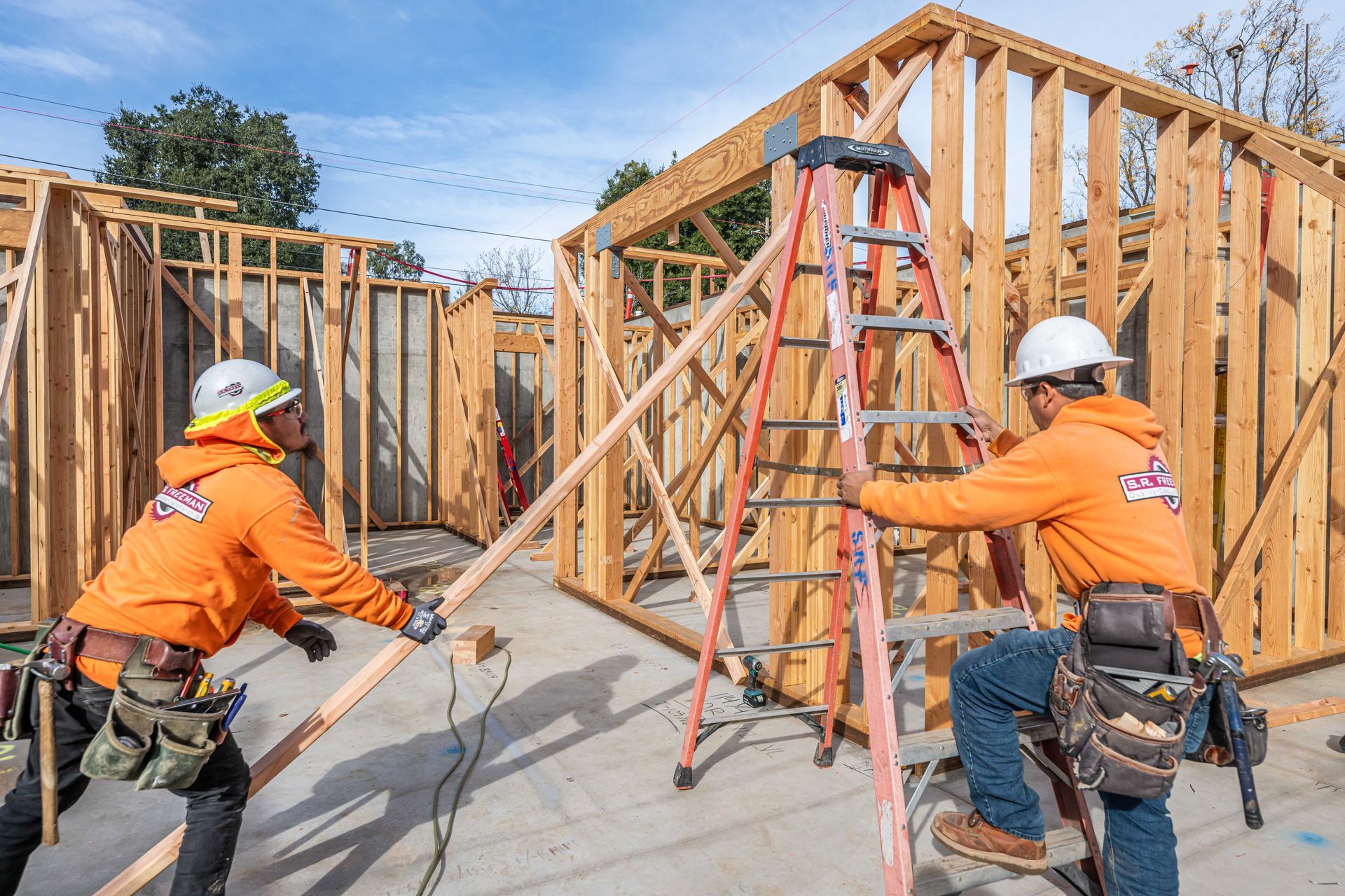 Construction workers framing a house, with ladders and wooden wall studs on a concrete foundation.