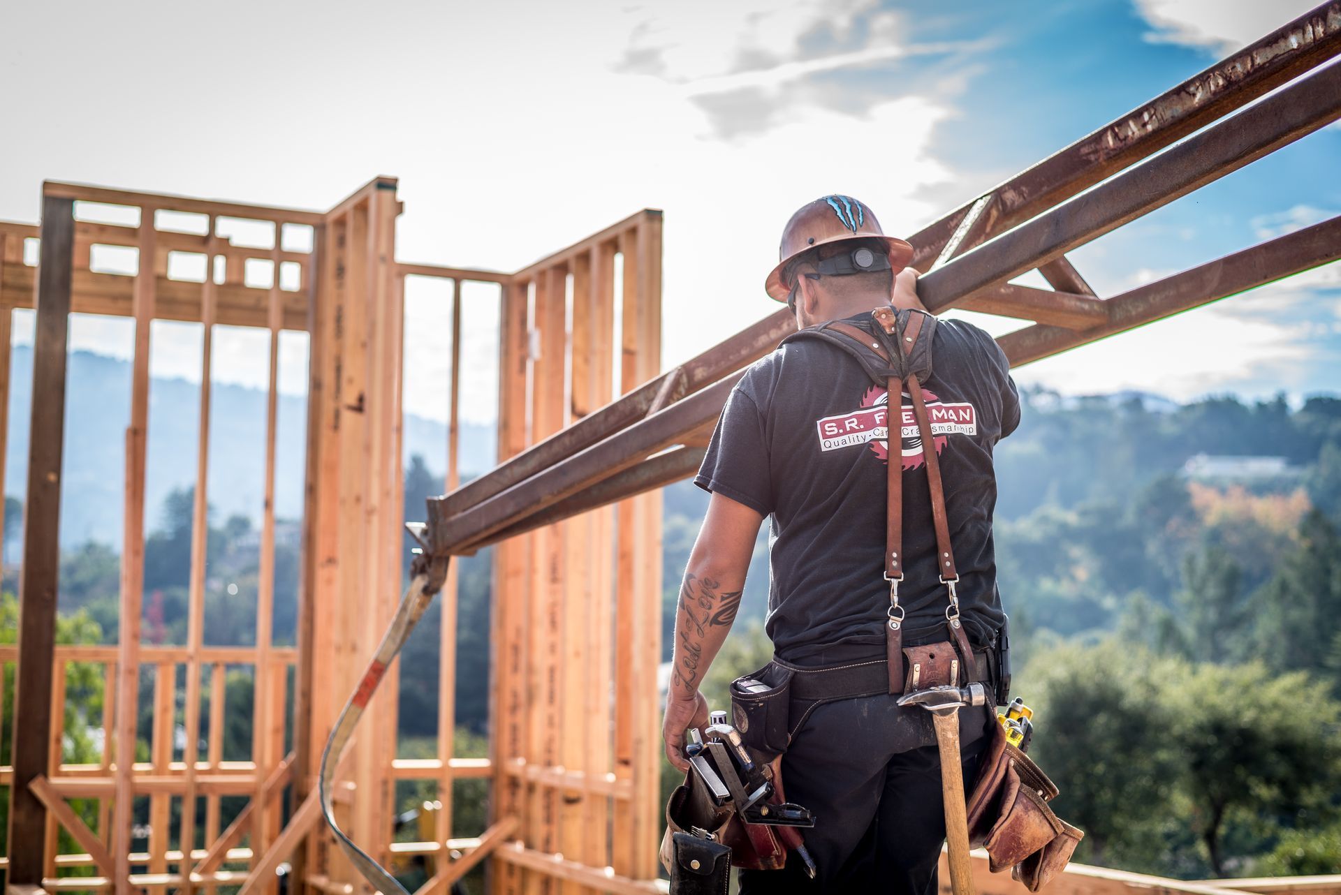 Construction worker on a wooden frame wearing safety gear, with mountains in the background.