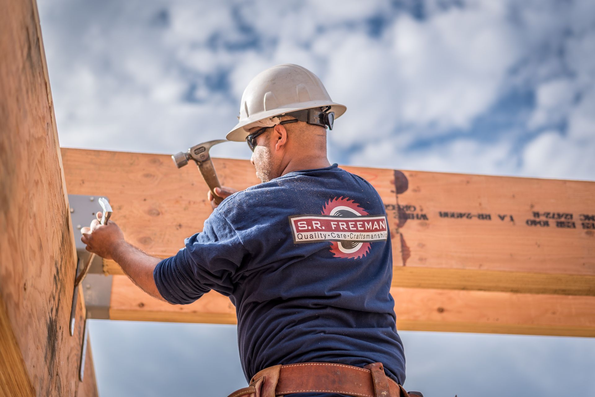 Construction worker hammering a wooden beam while standing on a frame under a cloudy sky