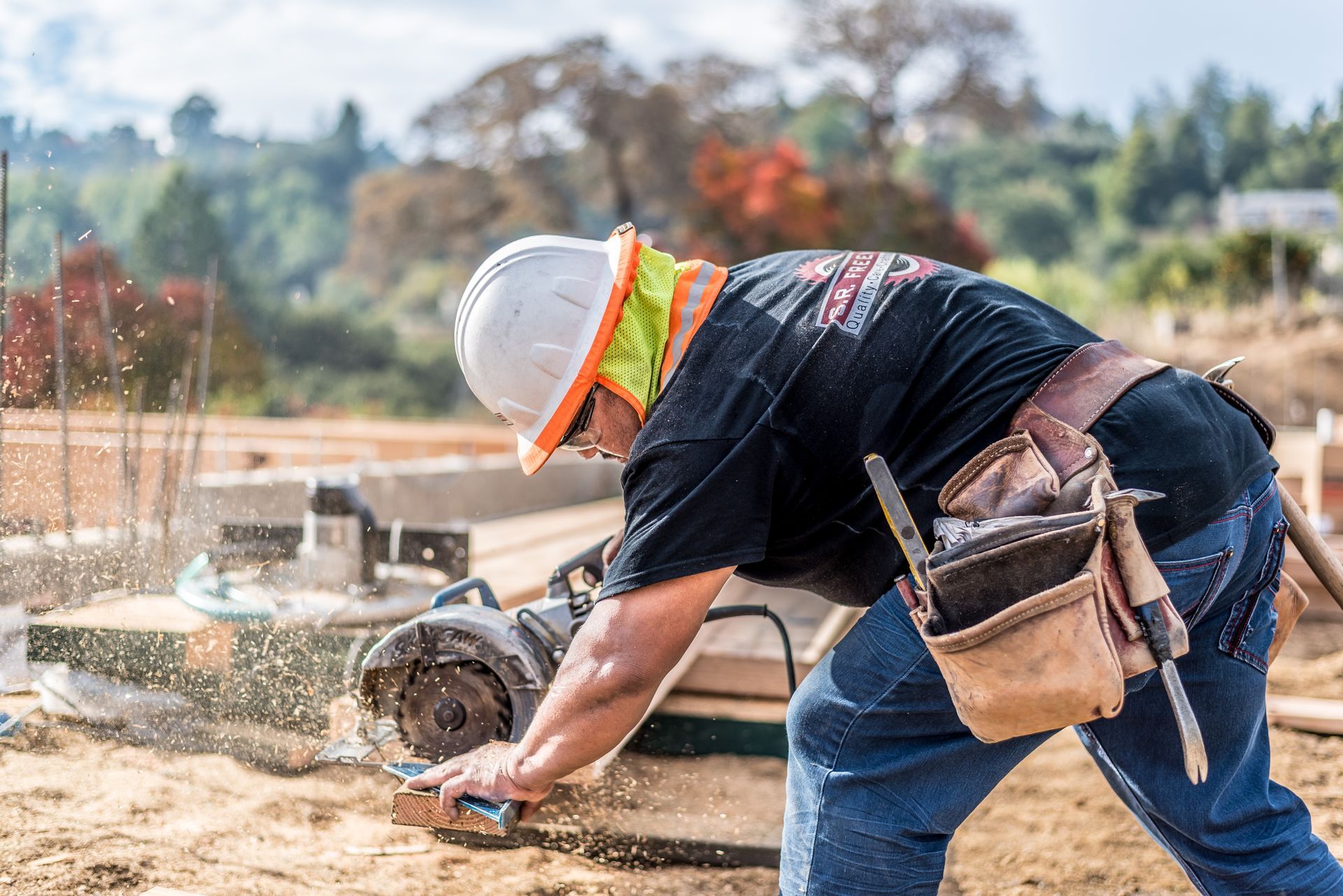 Worker in hard hat cuts concrete outdoors with a power saw, wearing safety gear and tool belt.