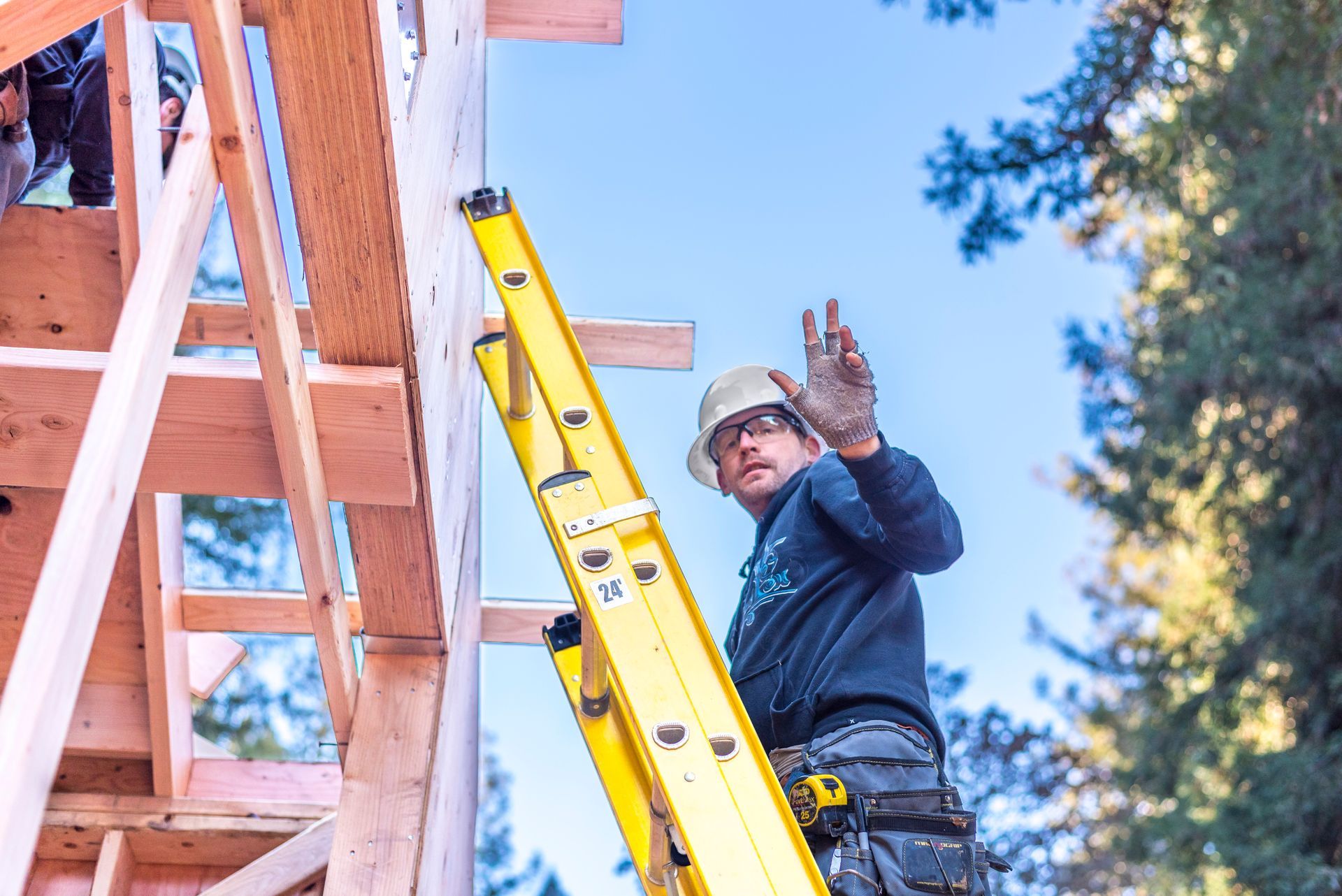 Worker on yellow ladder gesturing beside a wooden structure under construction outdoors
