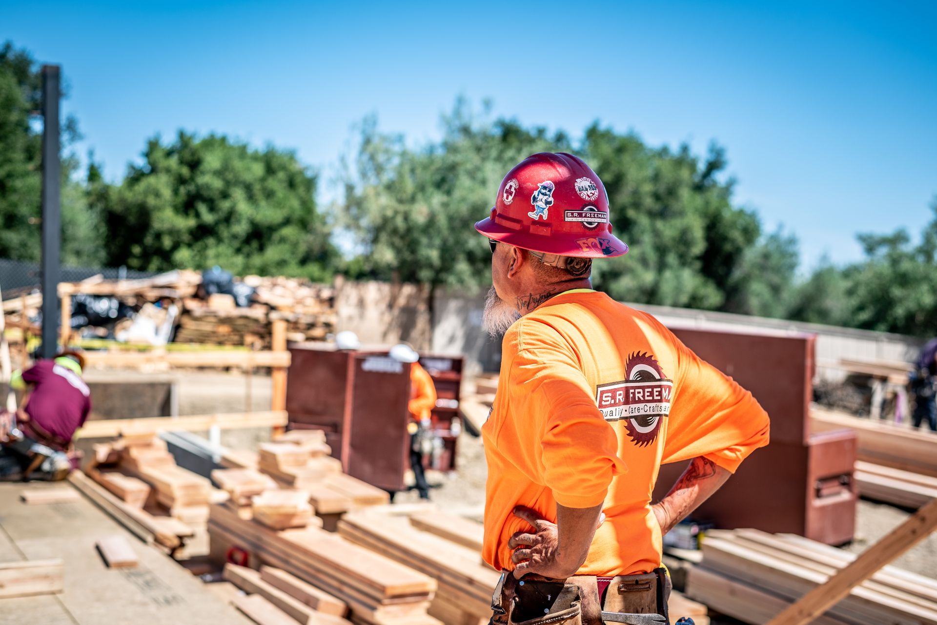Construction worker in an orange shirt and red hard hat stands at a building site with lumber and materials.