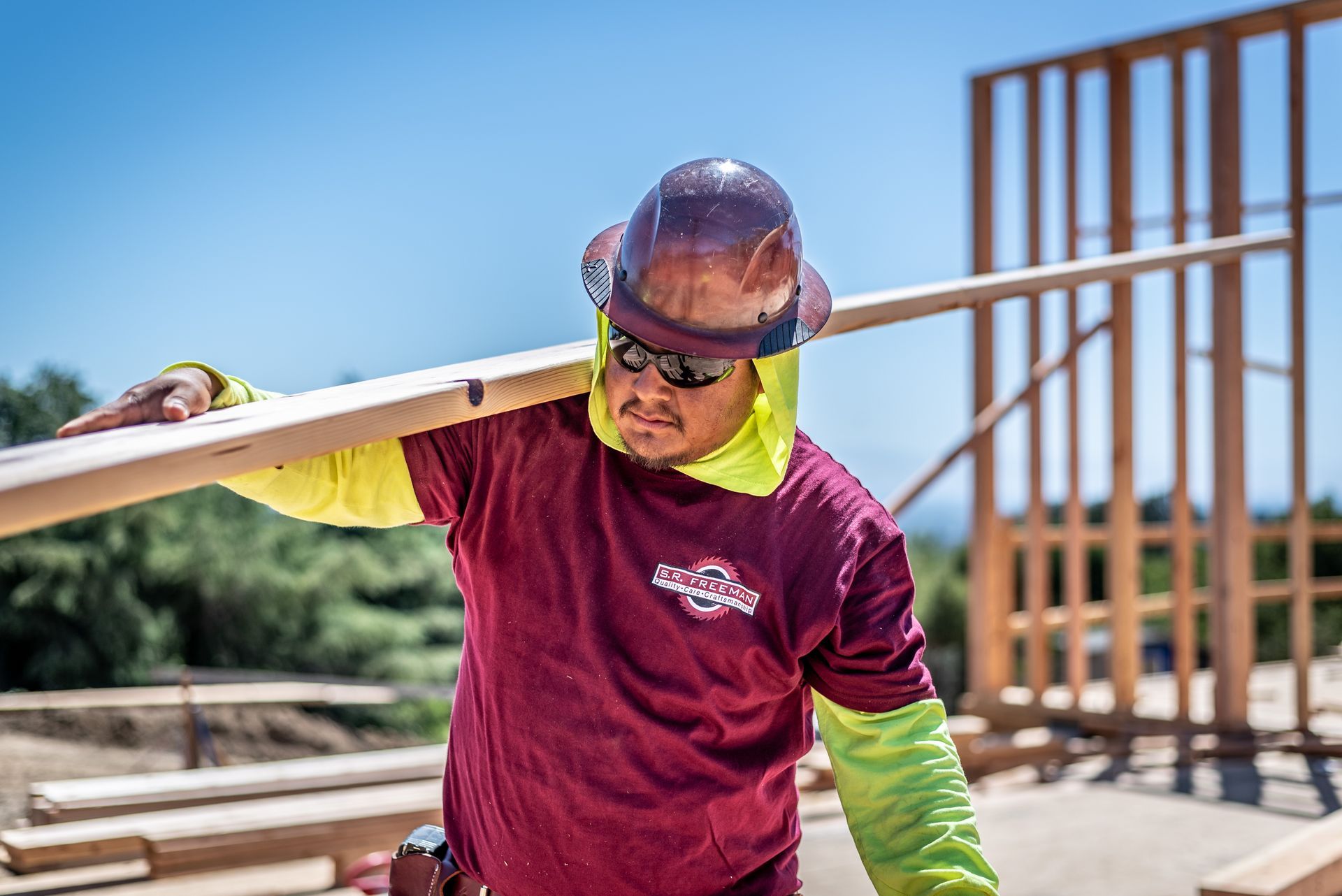 Construction worker in maroon shirt and hard hat carrying a beam at a building site