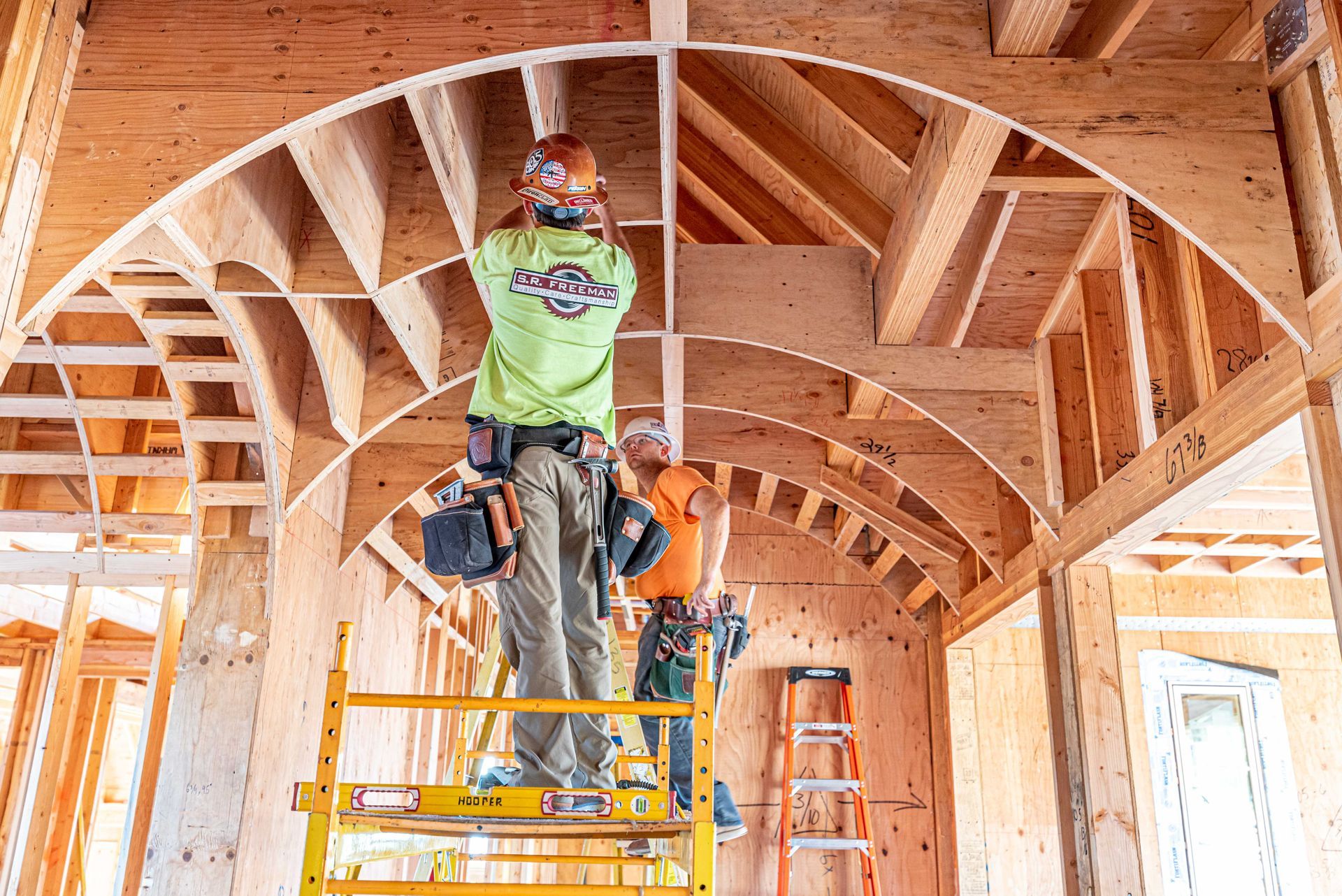 Construction worker on a ladder installing curved wooden beams inside a framed house.