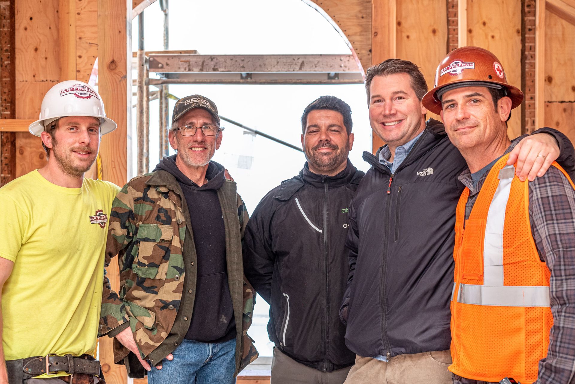 Five construction workers standing together indoors, wearing hard hats and safety gear, smiling at the camera.
