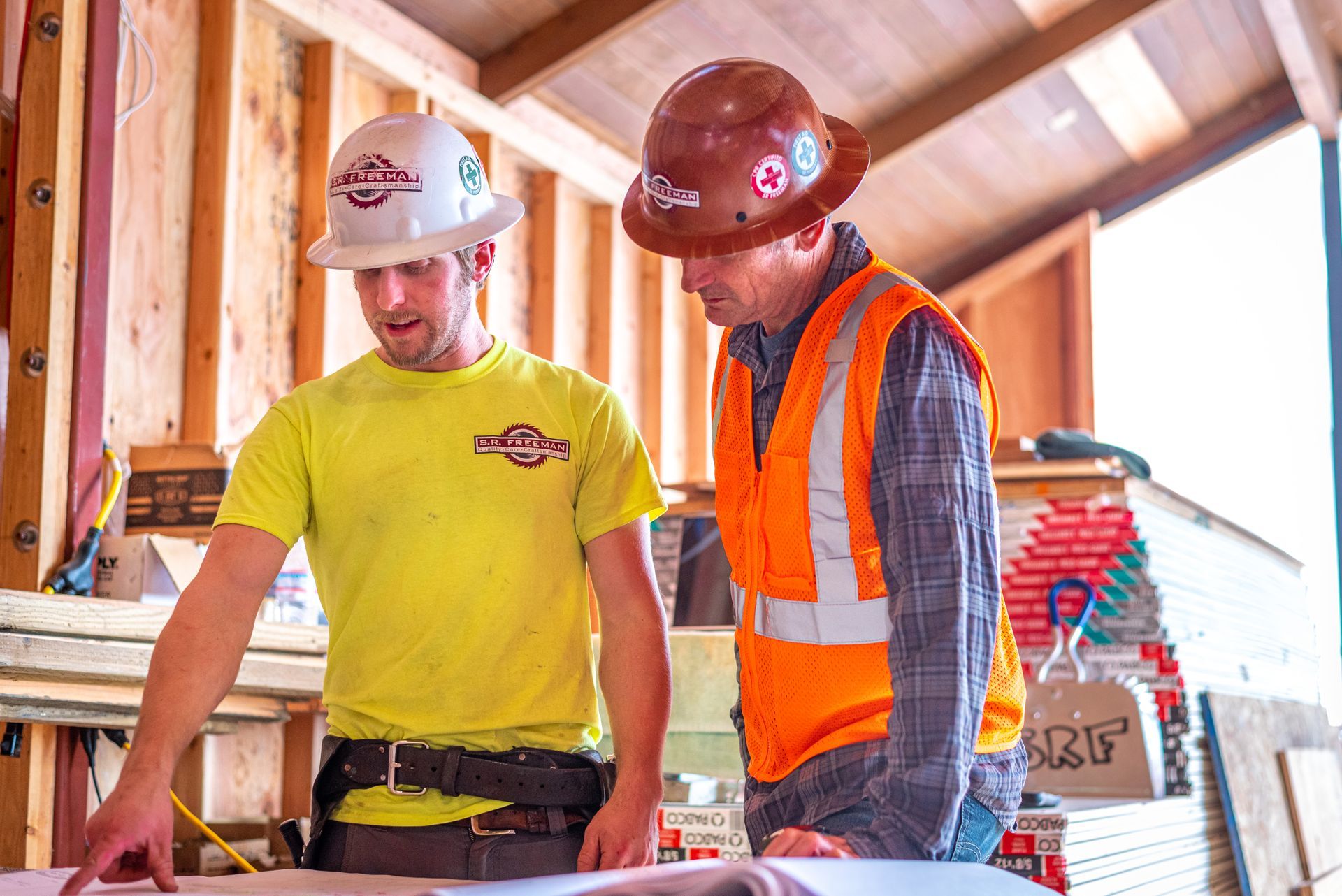 Two construction workers in hard hats reviewing plans inside a wooden-framed building under construction