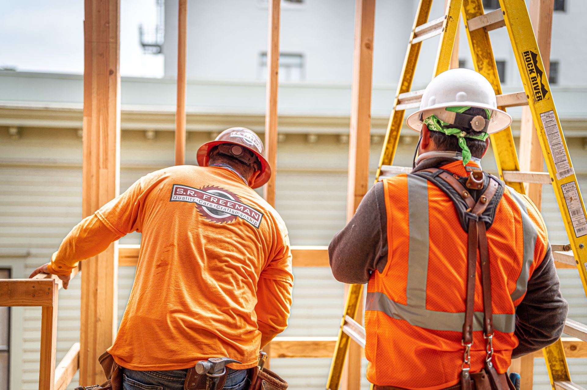 Two construction workers in orange safety vests and hard hats on a wooden frame indoors