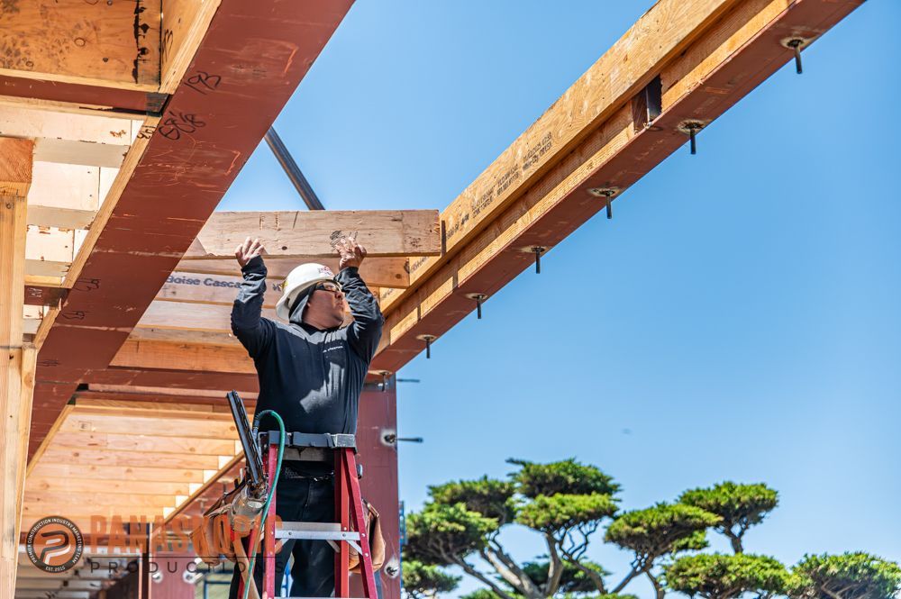 Worker on a ladder installing wooden beams against a clear blue sky.