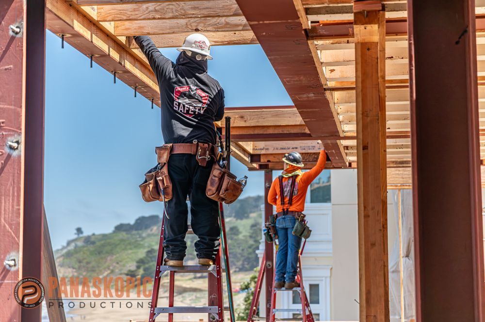 Workers on red ladders install a wooden ceiling frame at a construction site under blue sky