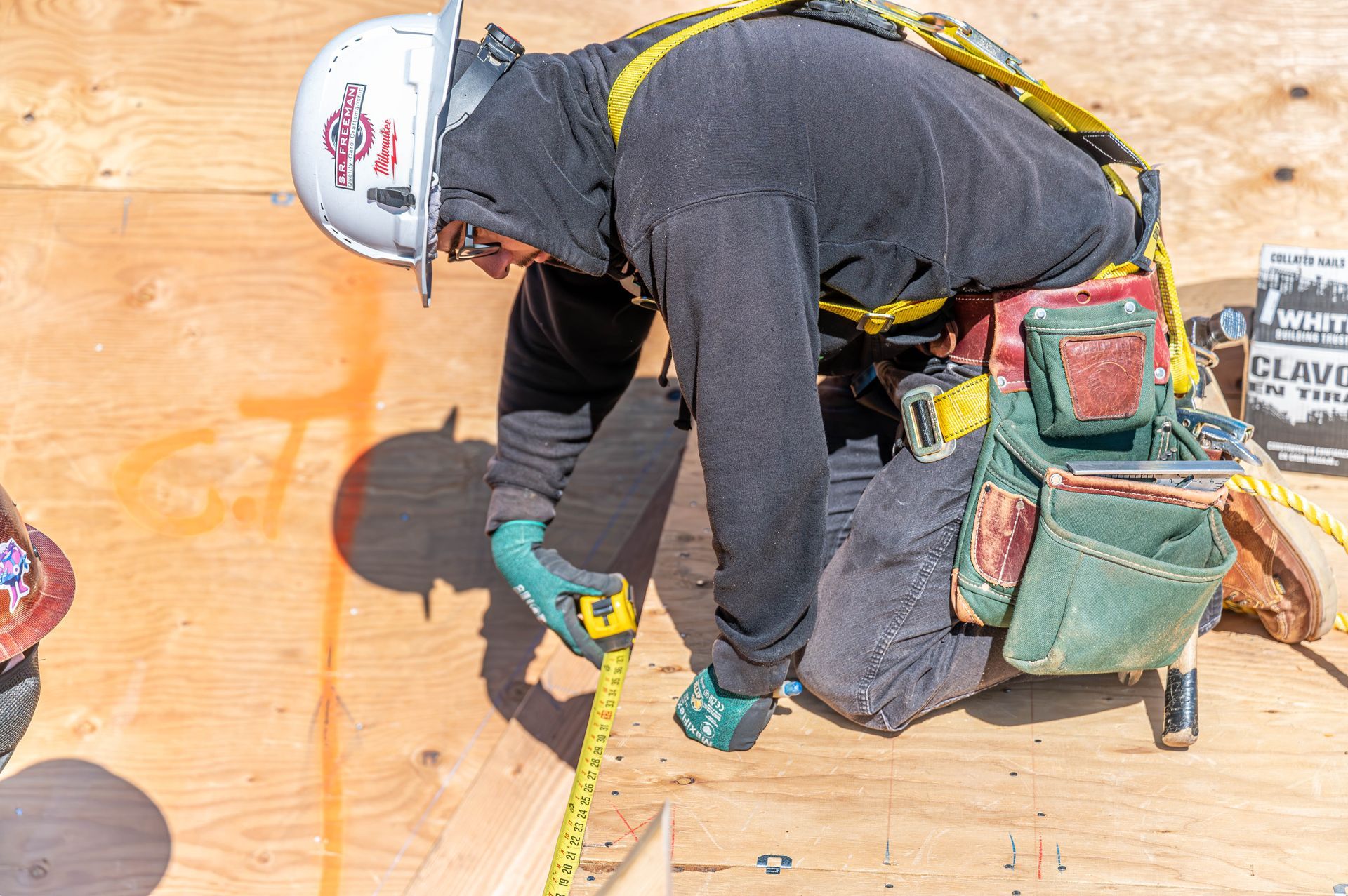 Construction worker kneels on plywood roof, measuring with a tape measure and wearing a hard hat and safety harness