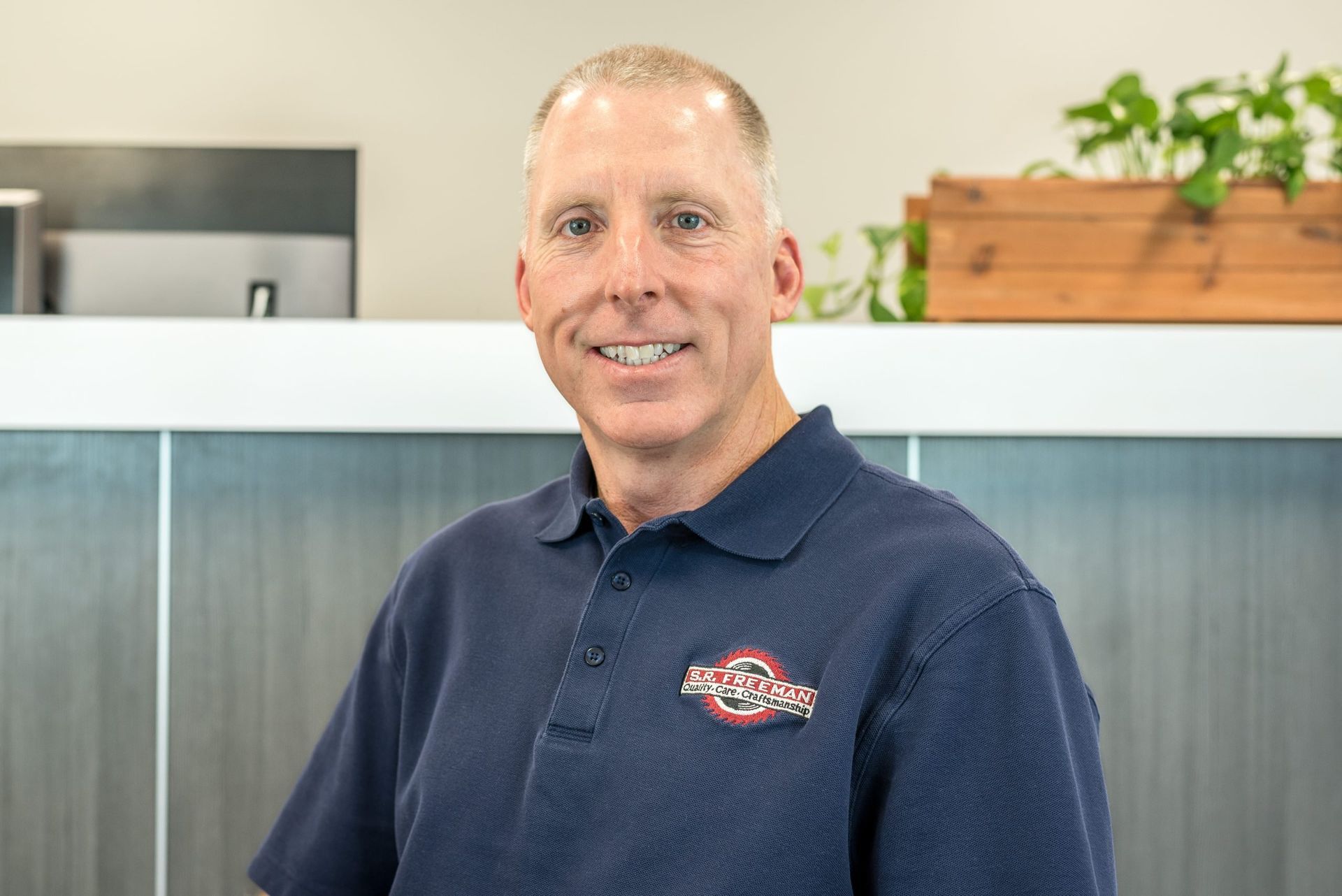 Man in navy polo smiling in an office lobby with white counter and plants in the background