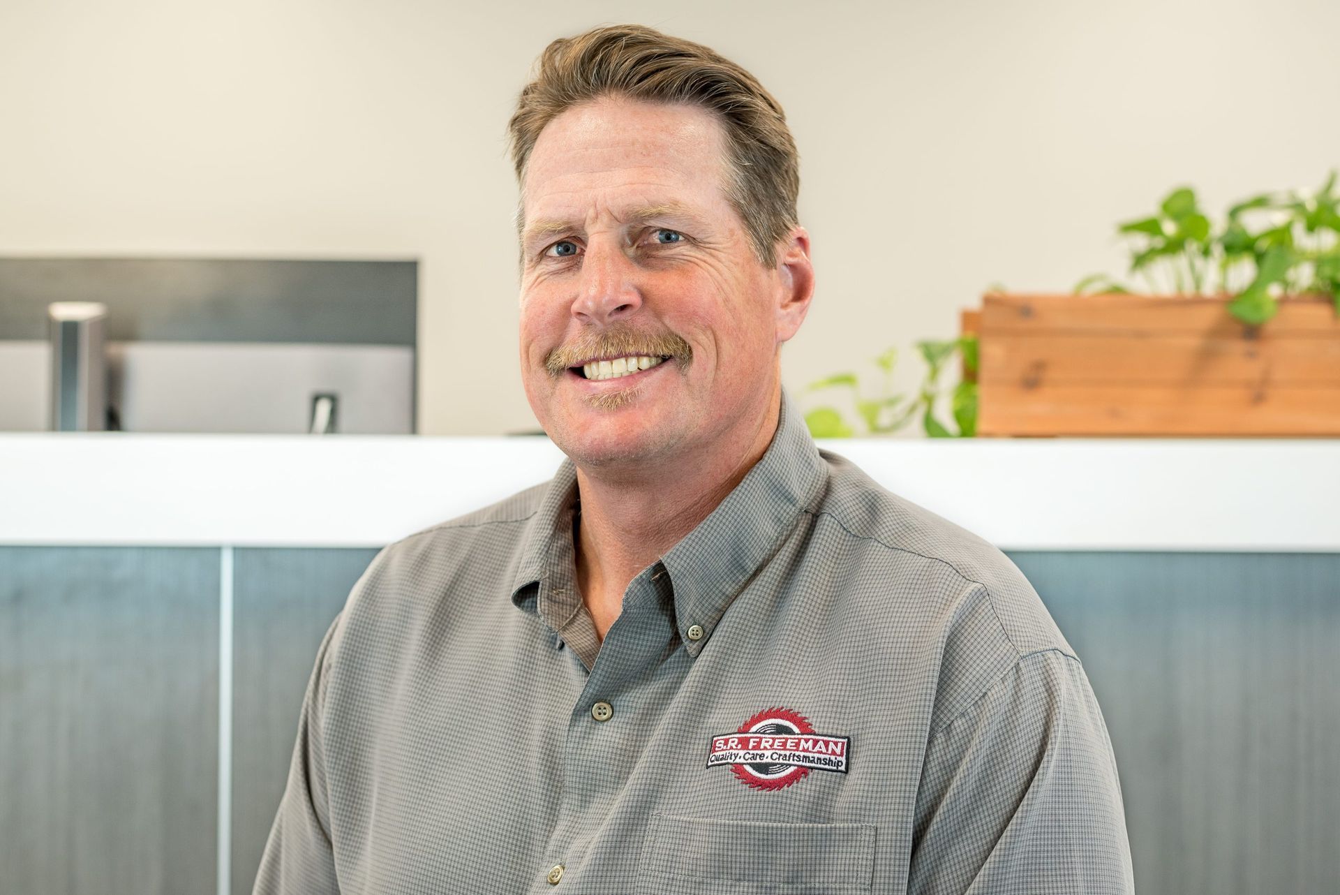 Man smiling in a gray button-up shirt standing in an office with frosted glass and a plant behind him
