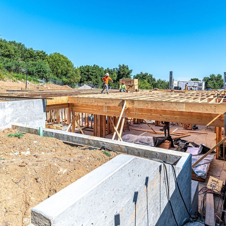 Construction site with concrete foundation walls, wooden framing, and workers under a bright blue sky.