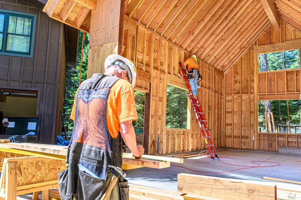 Workers framing a wooden interior, one standing on a ladder in a sunlit room.