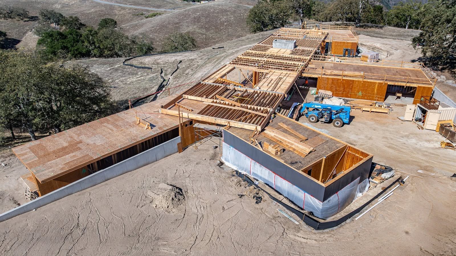 Aerial view of a partially built house with exposed wooden framing and orange sheathing on a dirt lot