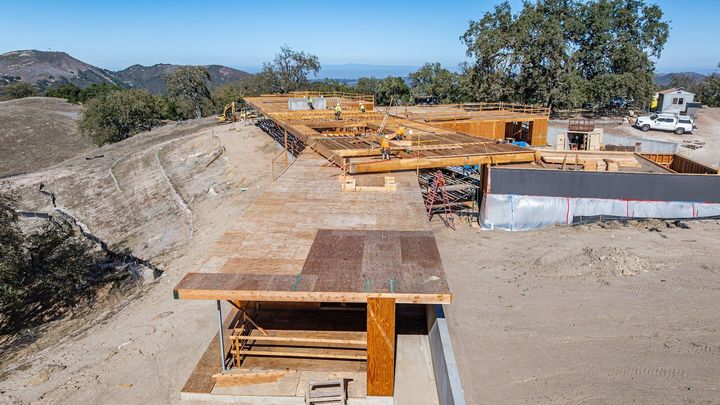 Construction site with wooden forms and concrete pads on a dry hillside, with trees and ocean in the distance