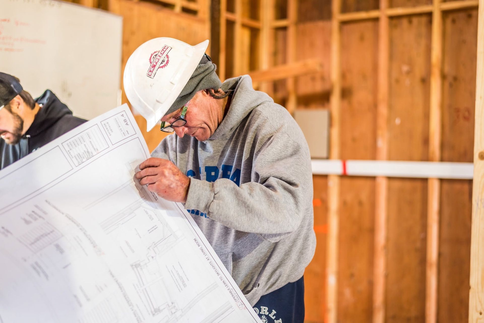 Worker inspecting construction plans in a wooden-framed building, wearing a hard hat and gray hoodie