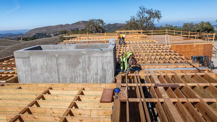 Construction workers framing a roof beside a concrete structure on a hillside under a clear blue sky