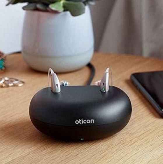 Audífonos Oticon en un estuche de carga negro sobre un escritorio de madera.