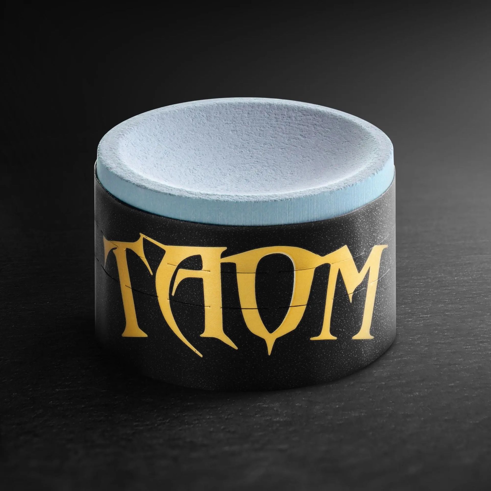 Black cylindrical candle with gold “TAOM” lettering and a pale blue top on a dark background