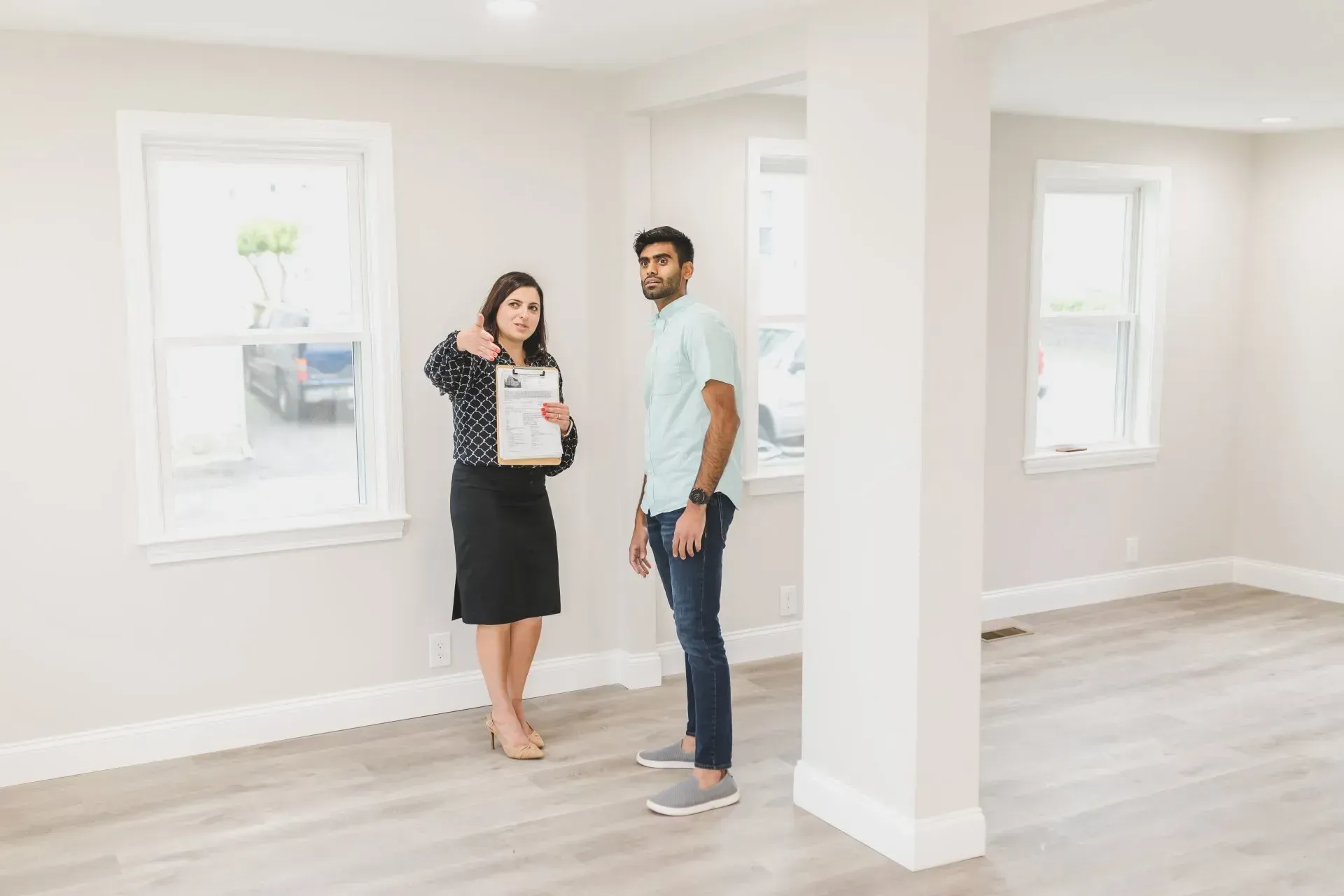Two people discussing a house in a bright, empty room with white walls and wood floors