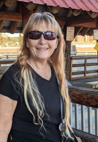 Smiling woman in sunglasses standing on a wooden deck under a red roof, with a railing and building behind her