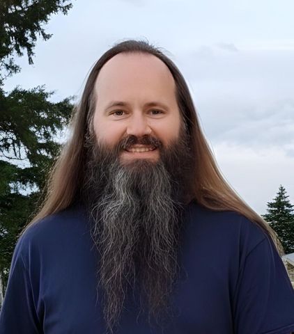 Smiling person with long brown hair and a gray beard outdoors under a cloudy sky.