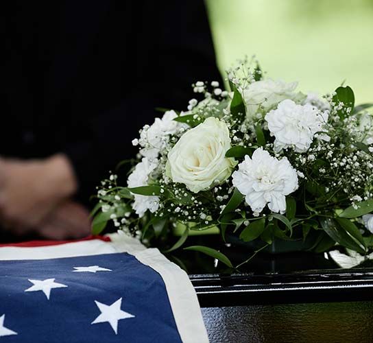 Black casket draped with an American flag, white flowers atop, person in black attire.