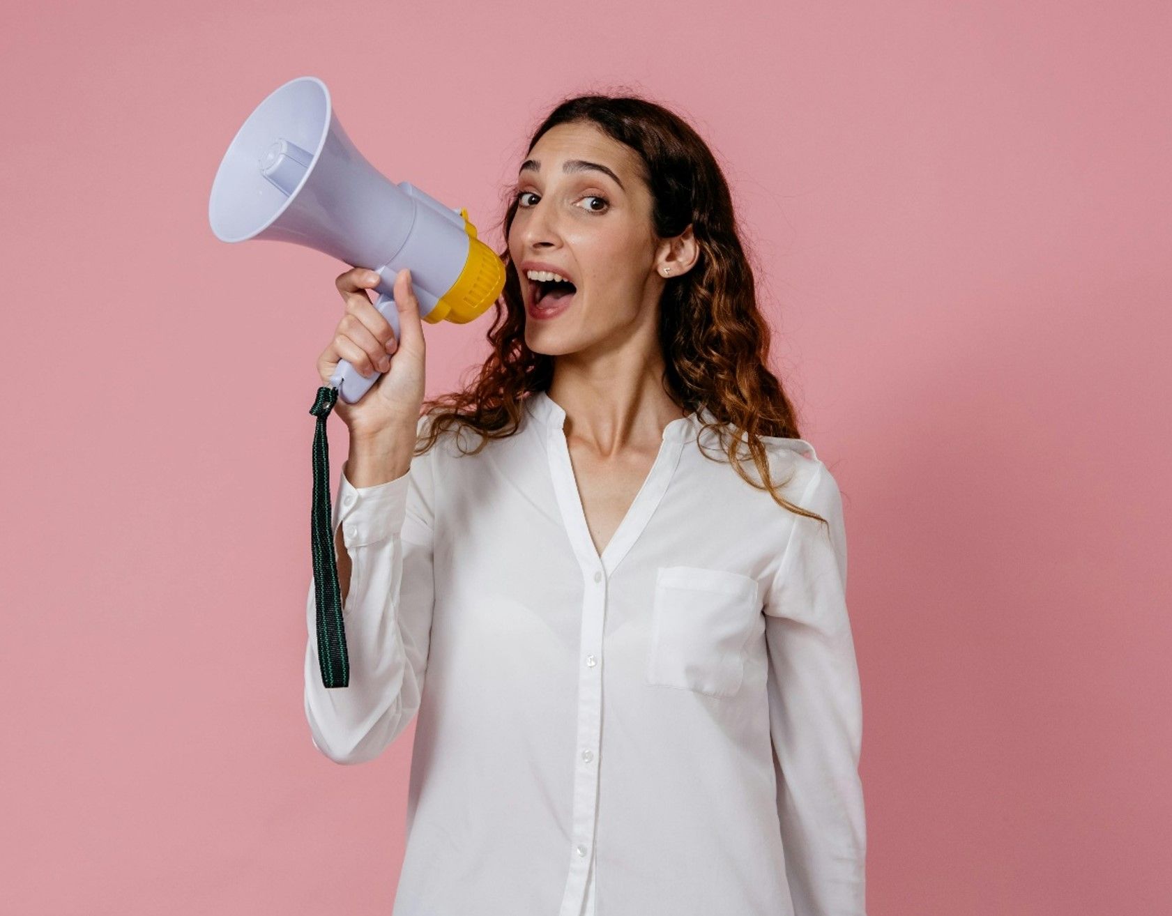 A person with long wavy hair, wearing a white shirt, holds a megaphone to their mouth against a solid pink background.
